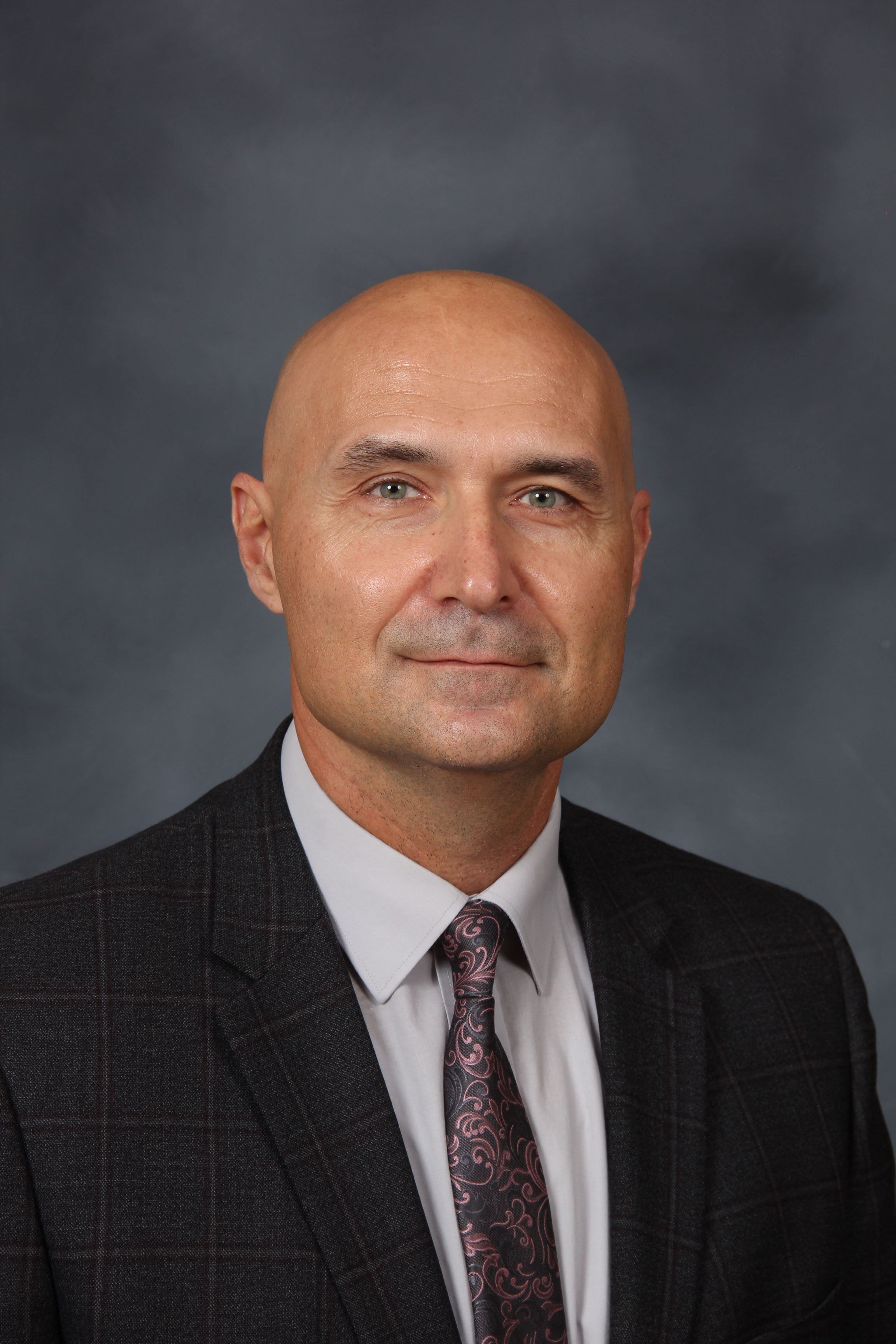 A man is wearing a suit and tie. He has a bald head and blue eyes. He is smiling and looking at the camera.