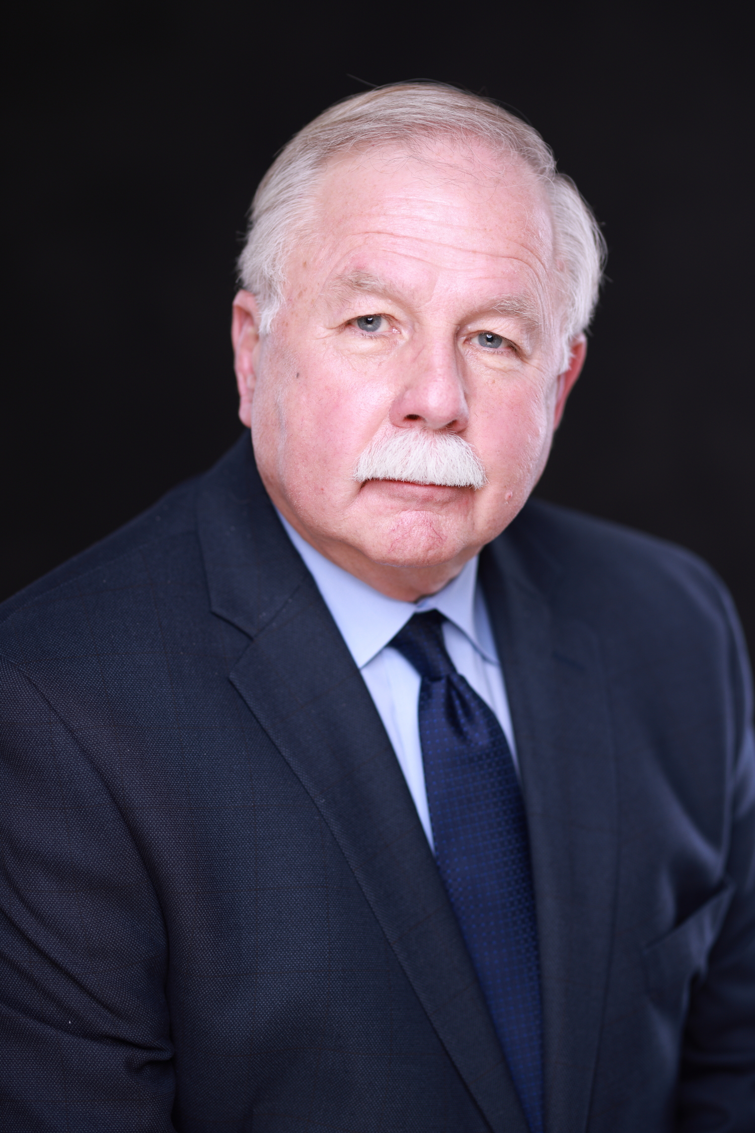 A man with a mustache is wearing a dark suit coat and tie.