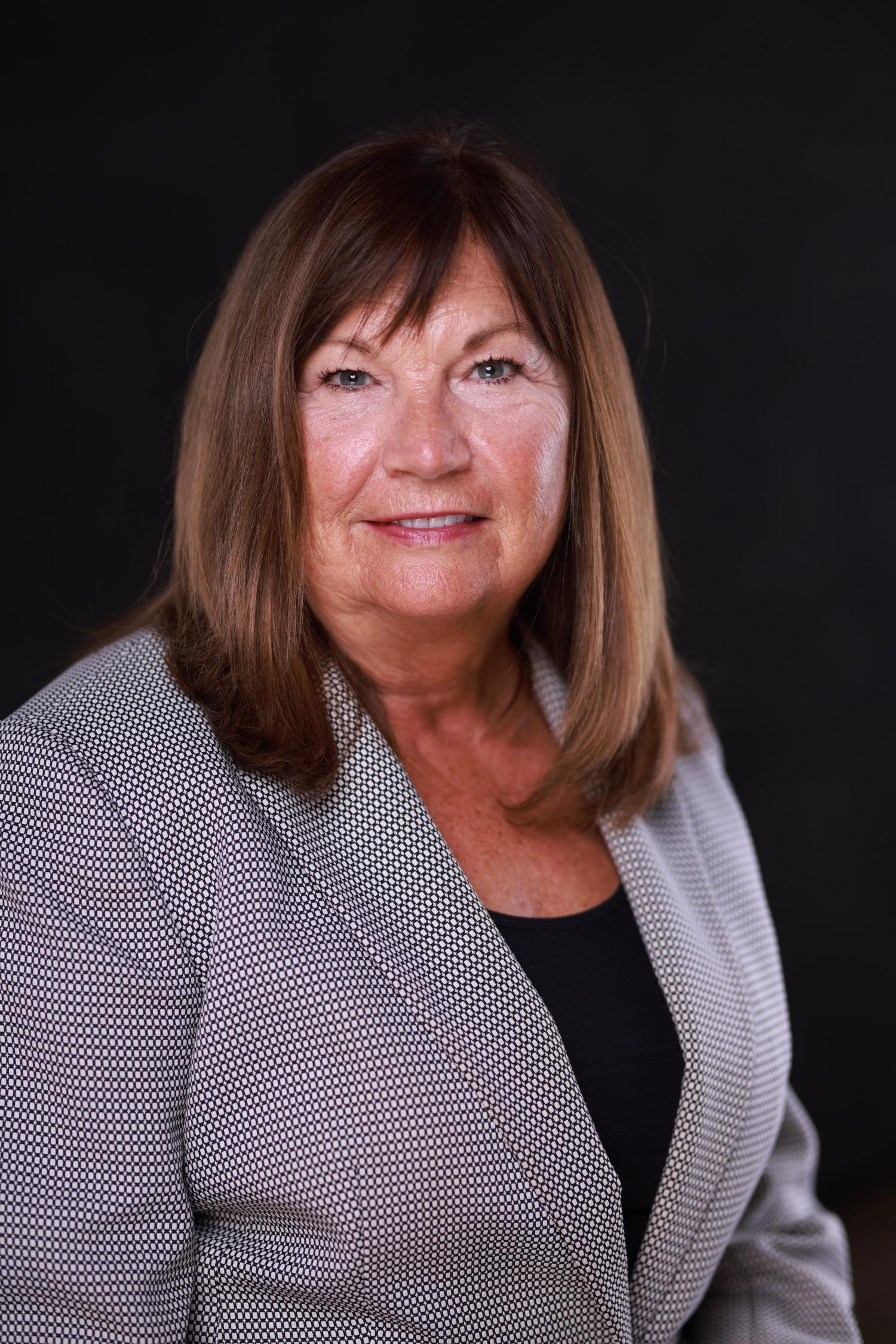 A woman with brown hair and a grey blazer is smiling.