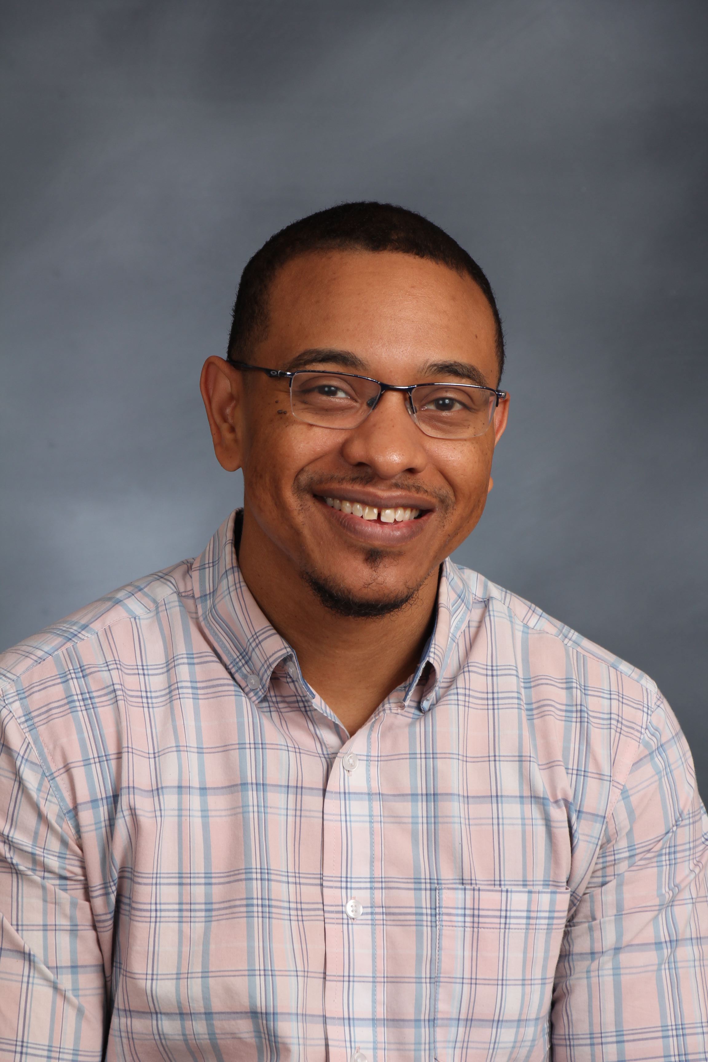 A headshot of a person wearing glasses and a pink and blue plaid shirt.