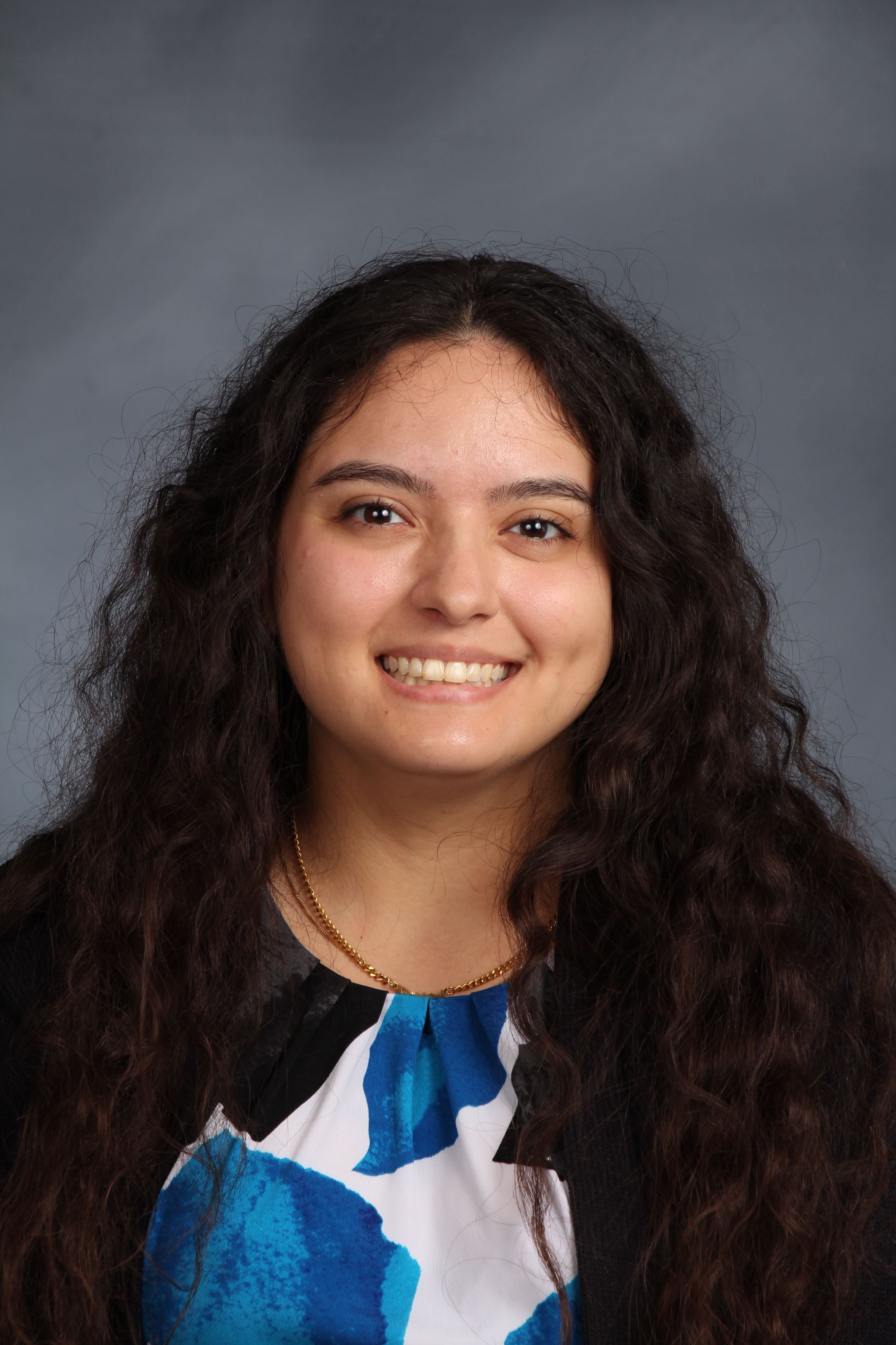 A headshot of a young woman with long brown hair. She is smiling and wearing a blue and white floral shirt.