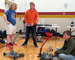Two boys stand and one sits as they operate two small robots on the gym floor at East Valley.
