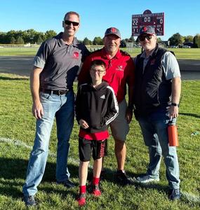 Shaun Roberts, in a red shirt, poses in front of the scoreboard with two school administrators and a young boy.
