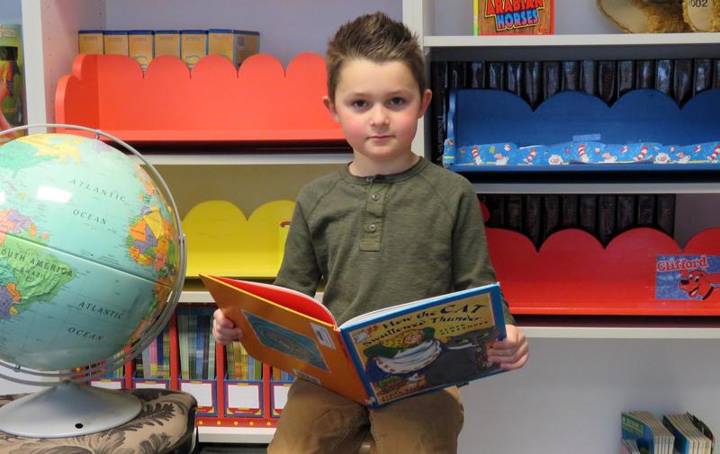 Kindergarten boys reads a book, surrounded by bookshelves and a globe.