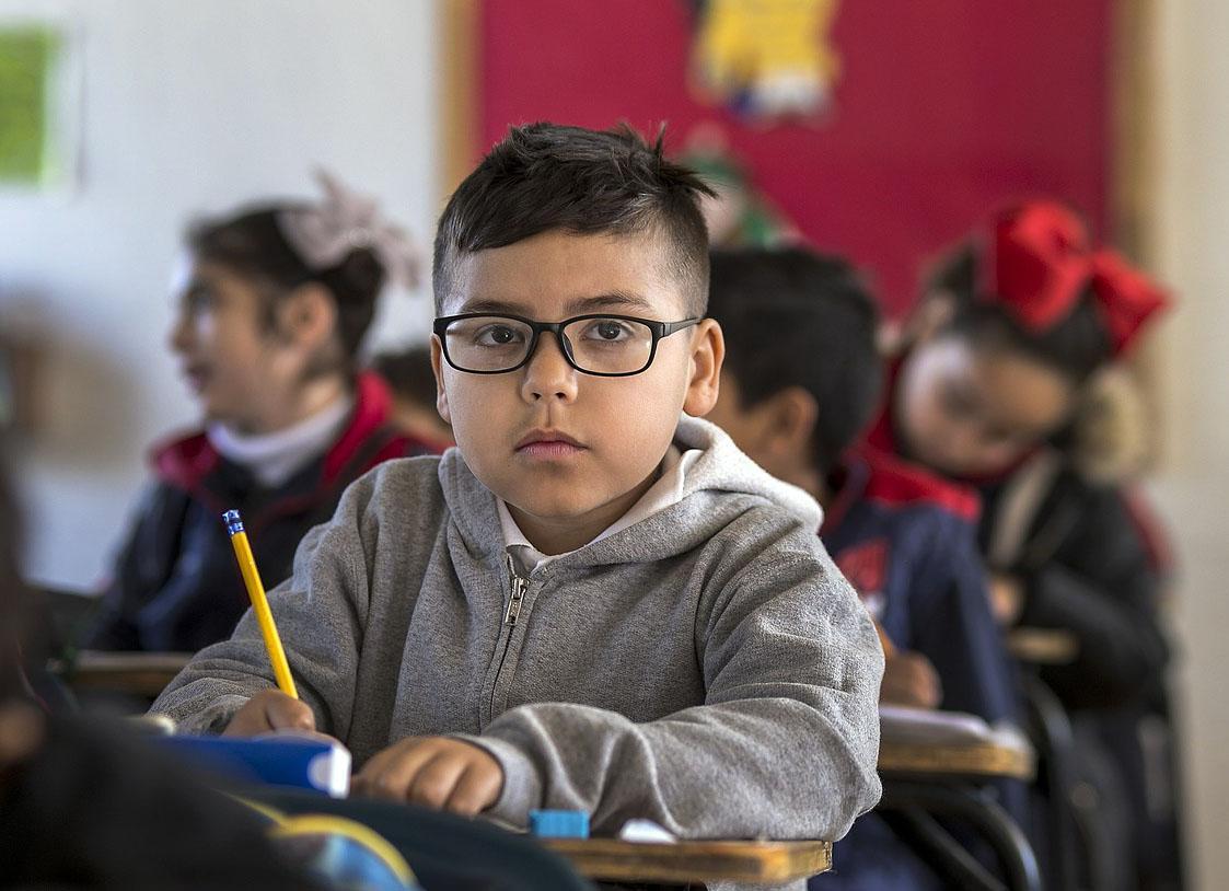 Boy with glasses holds pencil while sitting at a single desk.