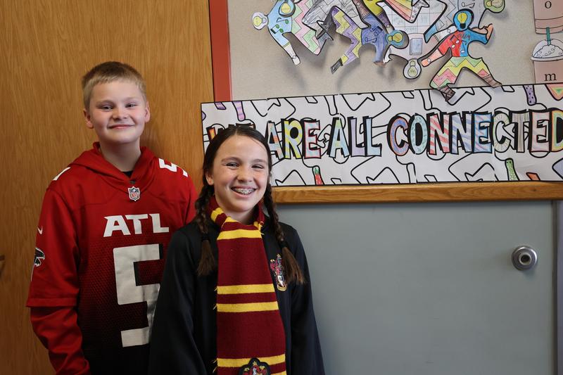 Boy in red sports jersey and girl in Gryffindor scarf pose in front of \"We are all connect\" sign.