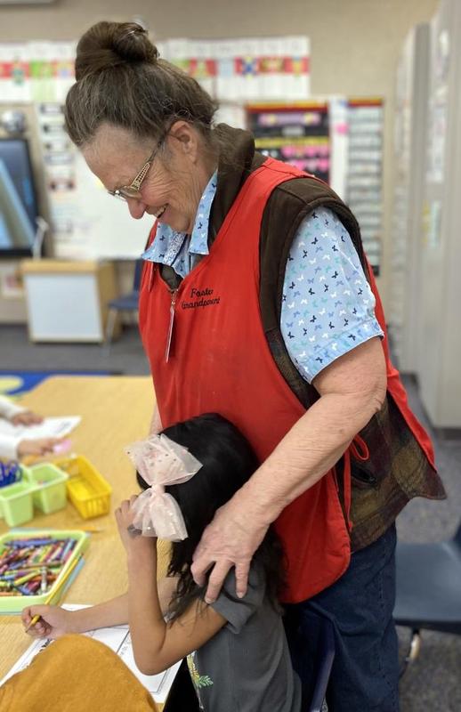 Senior woman in red vest squeezes shoulder of a dark-haired girl while looking at her worksheet.