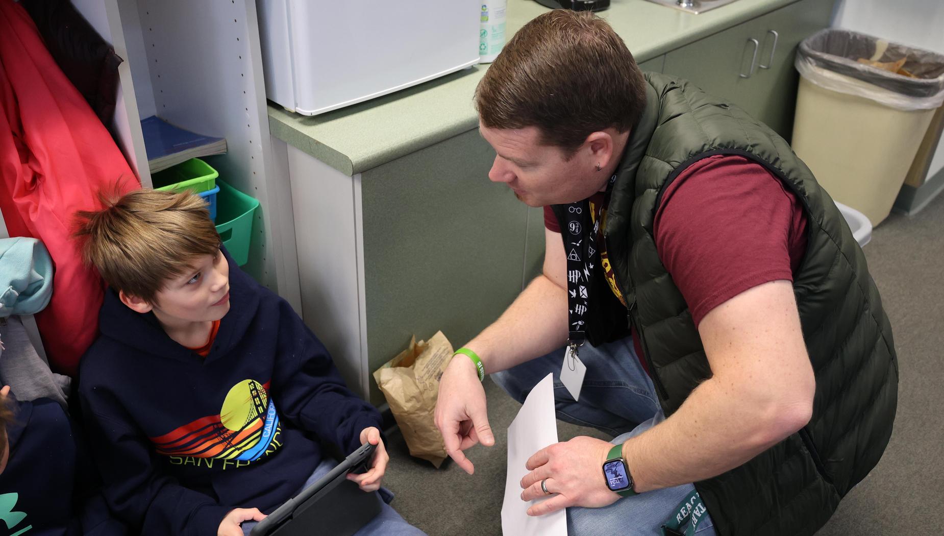 Principal squats down to talk to boy working on iPad on the floor.