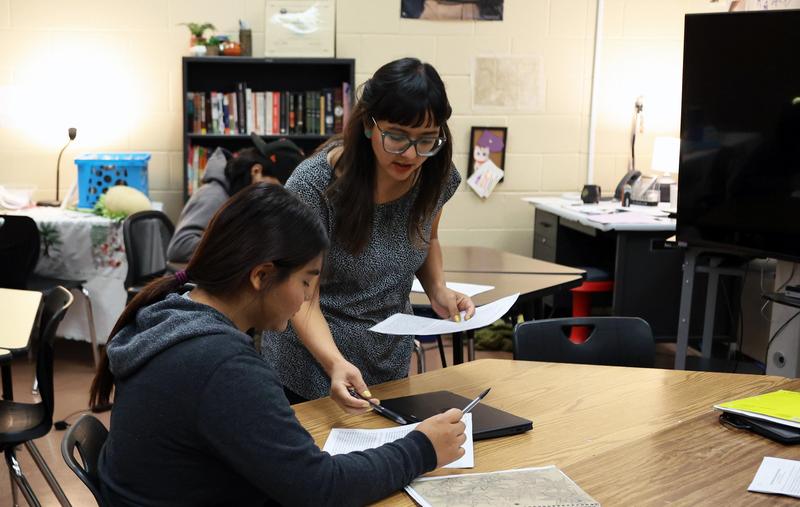 Teacher leans in to female student at work table to point out something on a paper.