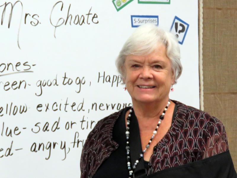 Silver-haired woman stands in front of whiteboard with  writing on it.