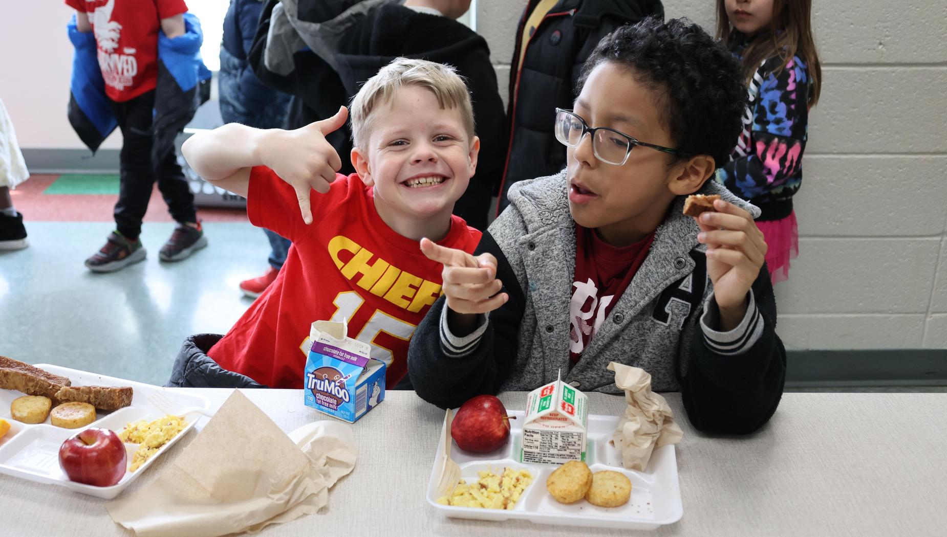 Boy in Chiefs T-shirt sits at the lunch table with arm draped around boy in A's jacket.