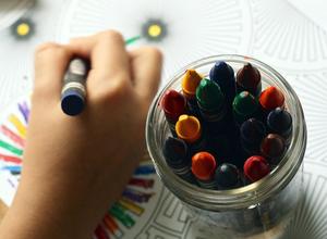 child\'s hand colors with a crayon, next to a full jar of crayons.