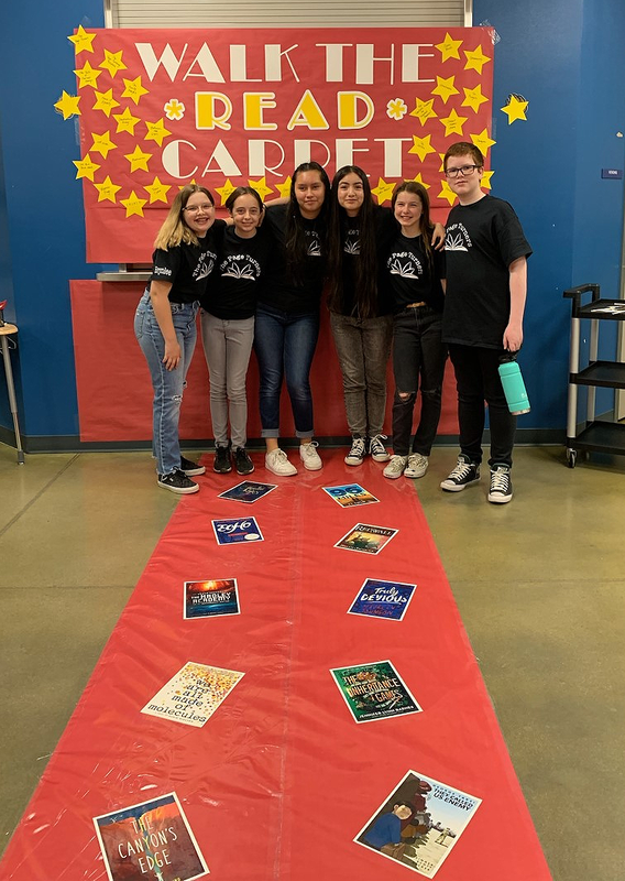 6 team members in club t-shirts pose on a \"read\" carpet decorated with cook covers.