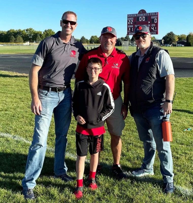 Shaun Roberts, in a red shirt, poses in front of the scoreboard with two school administrators and a young boy.

