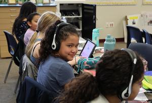 Girls in blue shirt and white headphones turns to look at camera while working on her computer in class.