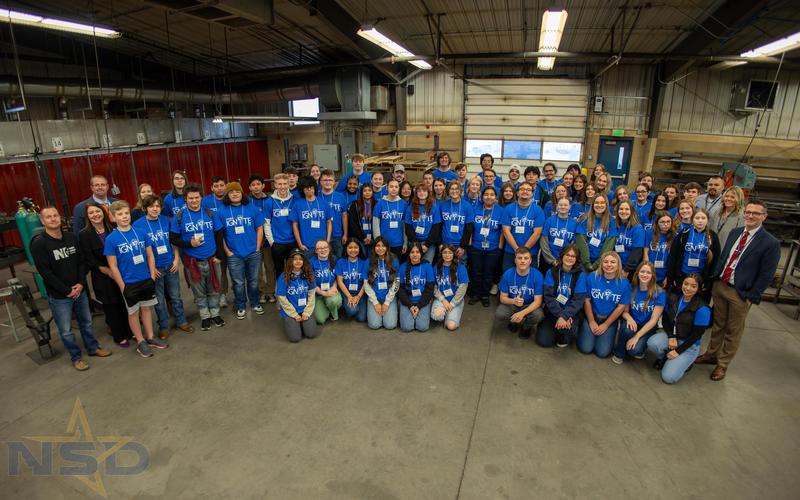 Students in Nampa IGNITE blue T-shirts pose in semicircle with adult organizers.