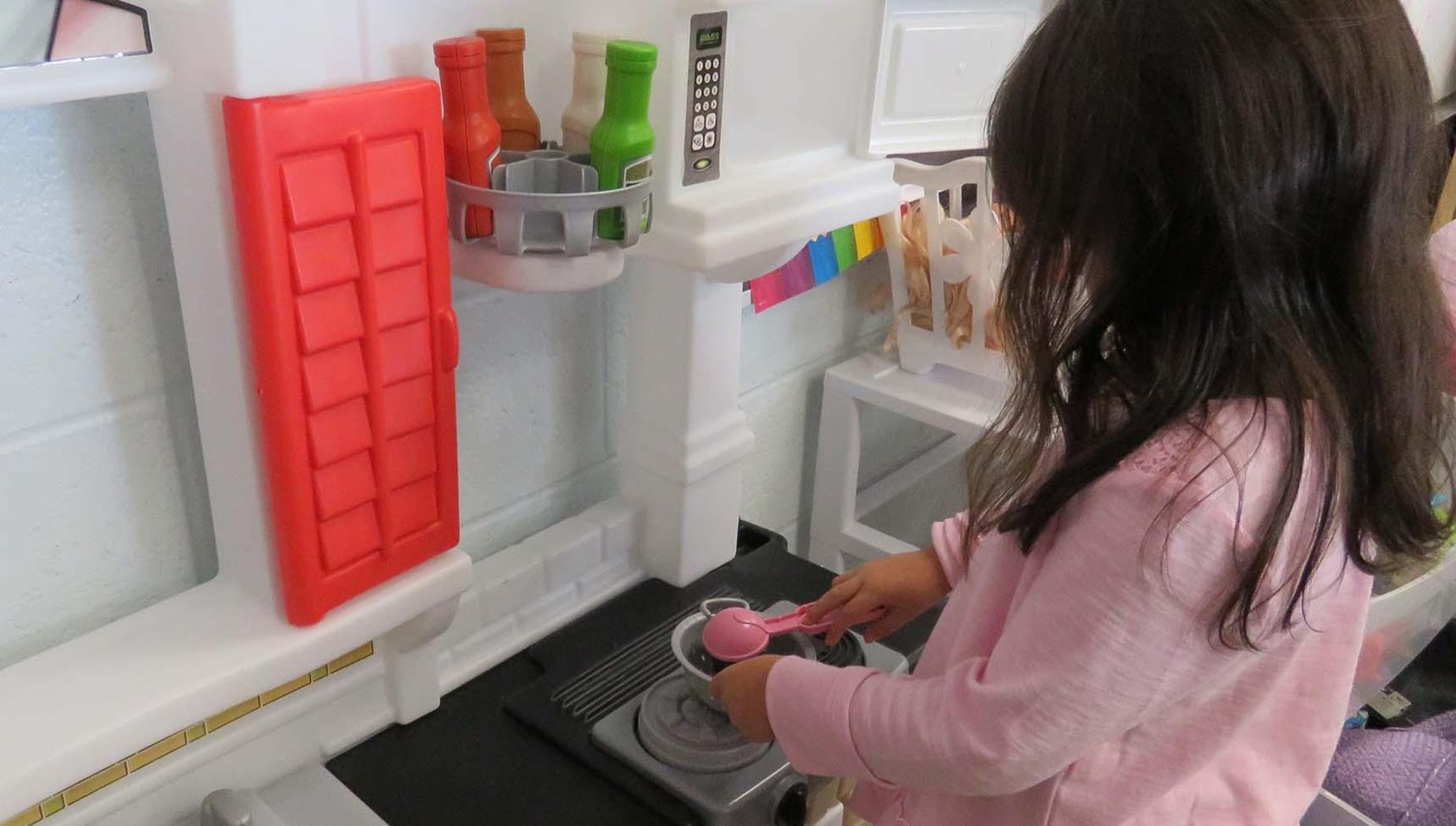 A young girl dressed in pink pretends to cook in a play kitchen.