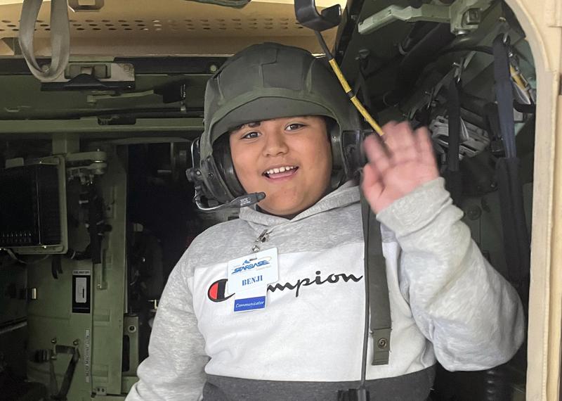 Young boy wearing a helmet with a mic waves from the doorway of a military transport vehicle at Gowen Field.