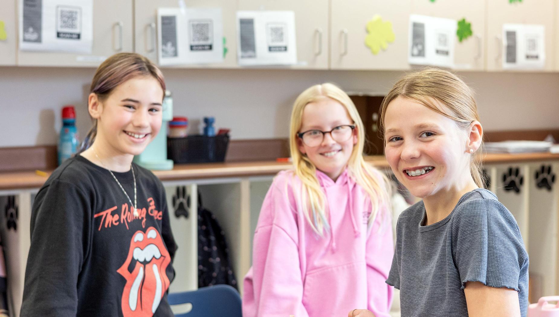 Three girls, dressed in black, pink and gray, smile at the camera in the classroom.