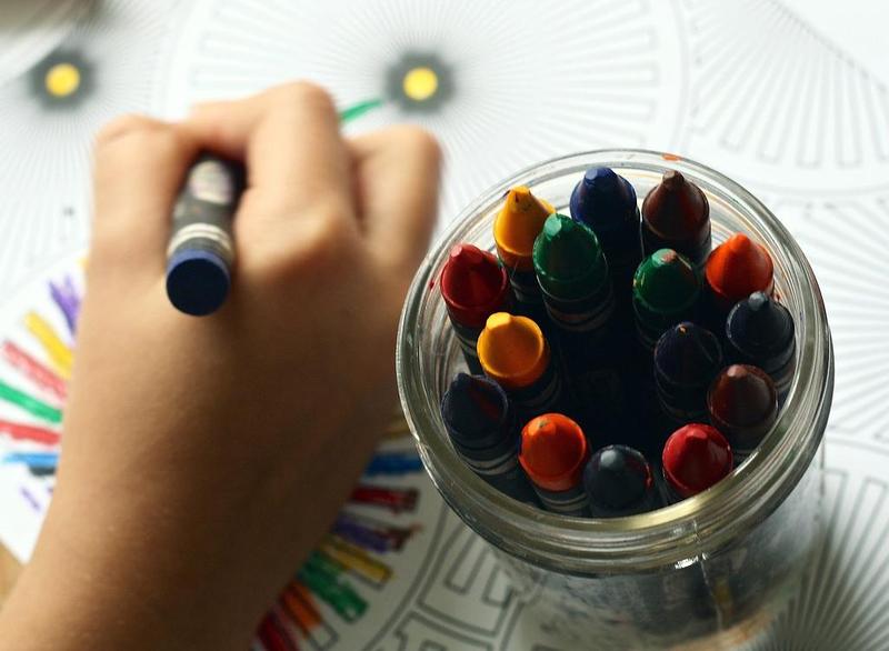 child\'s hand colors with a crayon, next to a full jar of crayons.