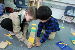 Two young boys kneel on the carpet almost touching heads as they arrange counting tiles.