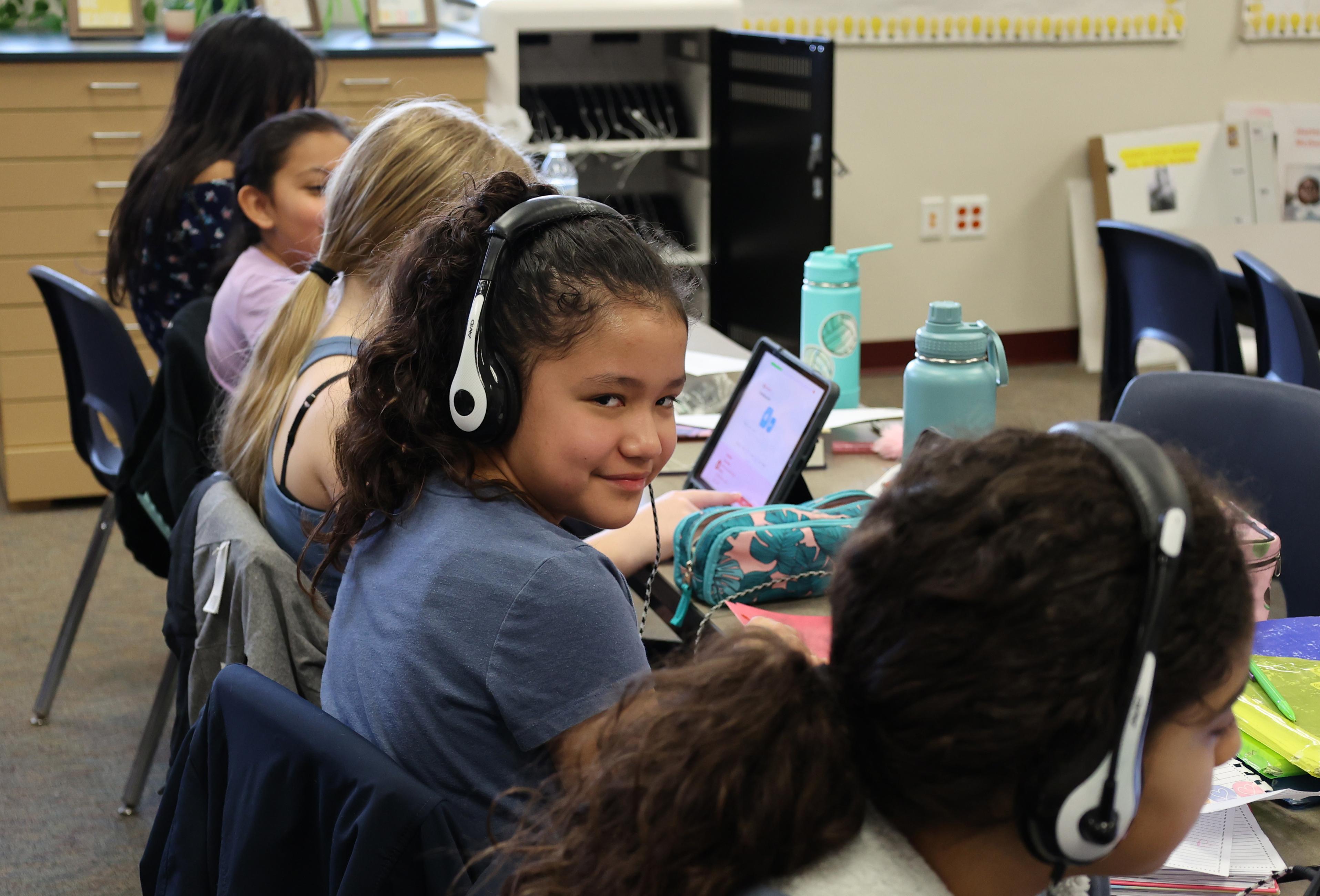 Girls in blue shirt and white headphones turns to look at camera while working on her computer in class.