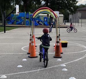 A child on a blue bike, wearing a red helmet, bikes through at tunnel made of bent pool noodles as part of a bike obstacle course.
