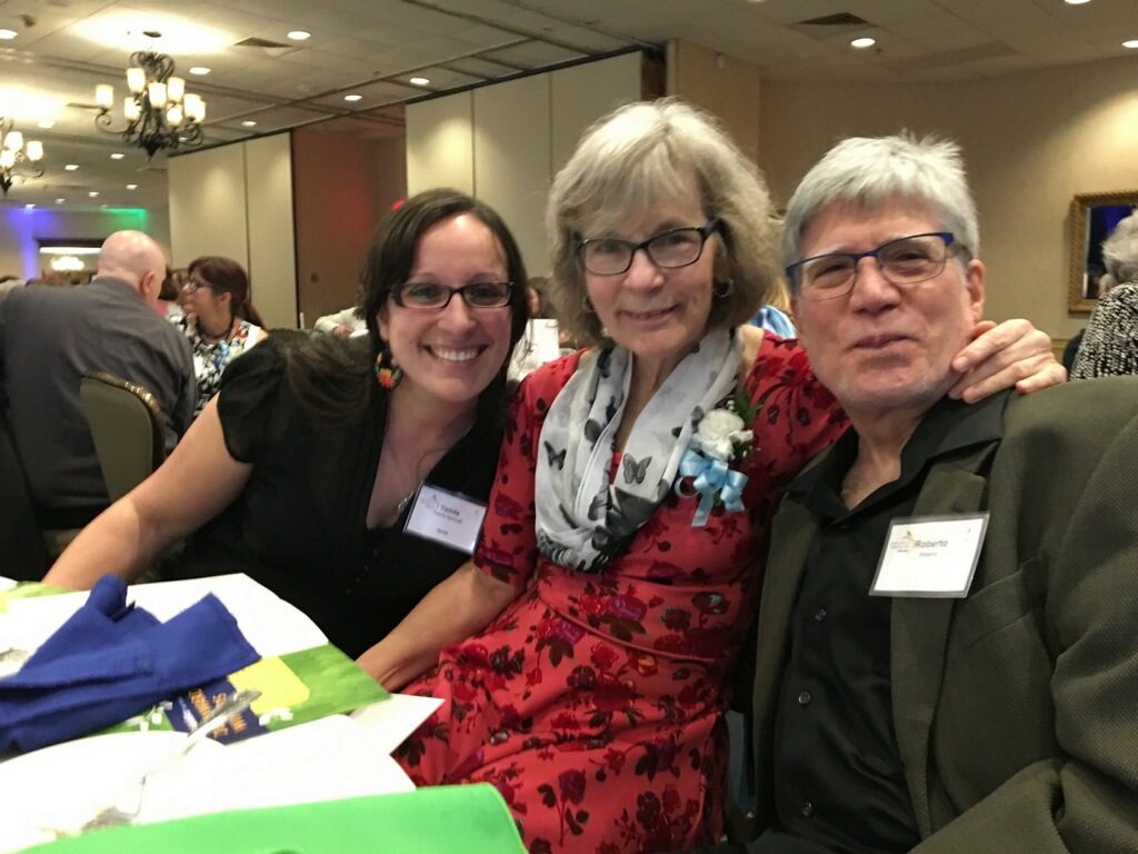 Three people sit together at the table facing the camera. Teacher Kim Gerould is the central figure.