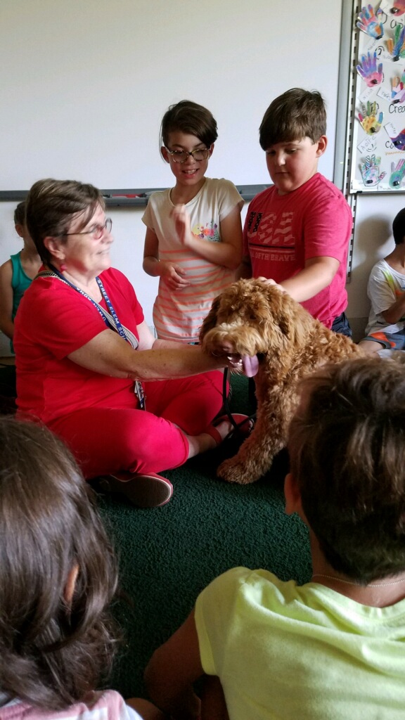 Principal Agna sits on the floor with Jackson the dog as two 3rd grade students pet Jackson.