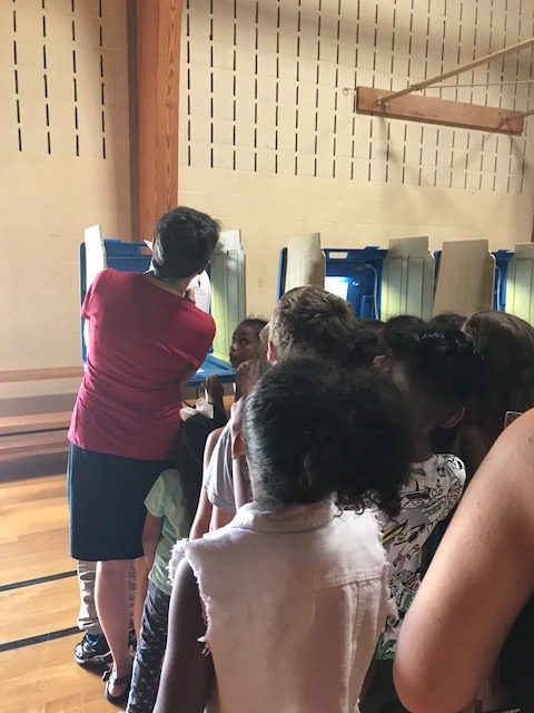 Teacher Mary Cowhey with back turned showing students voting ballot.