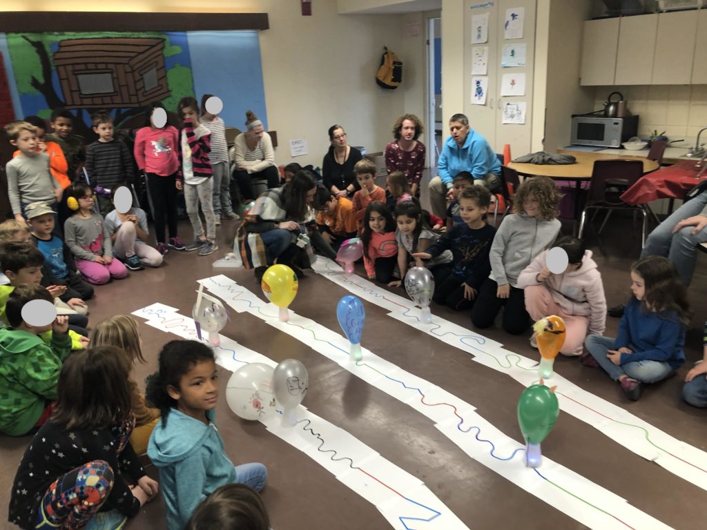 Students sit in a room watching small robots carry decorated plastic cups in a parade format.