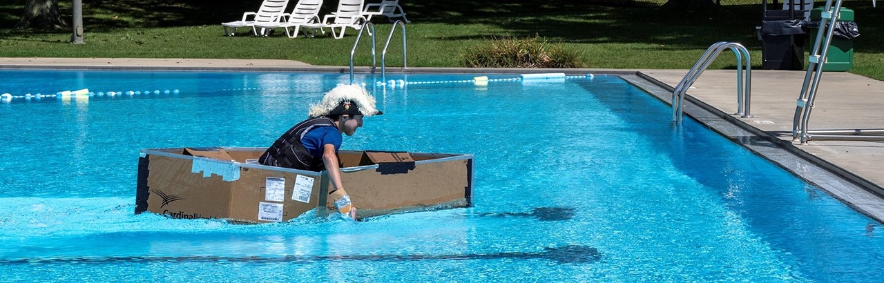 high school male student wearing an admiral hat paddles a cardboard boat in a swimming pool