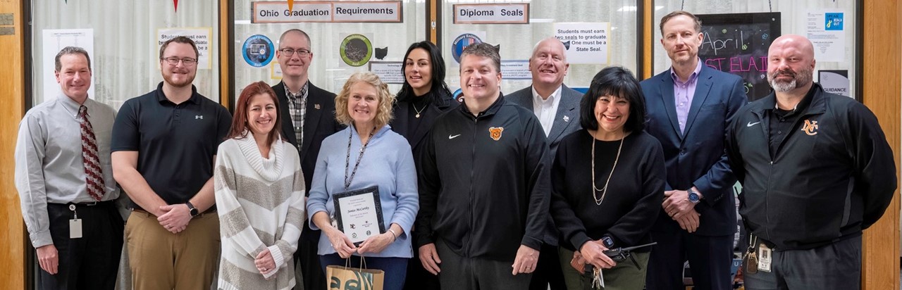 District and high school administration with community representatives honor blonde teacher in the middle of group