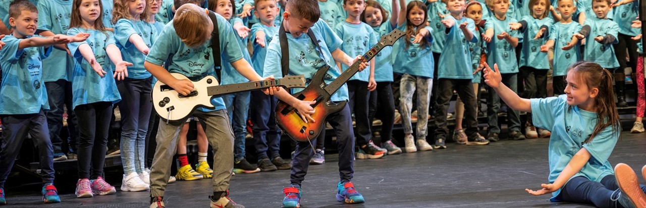 two boys pretending to play the guitar with second grade students behind them on risers