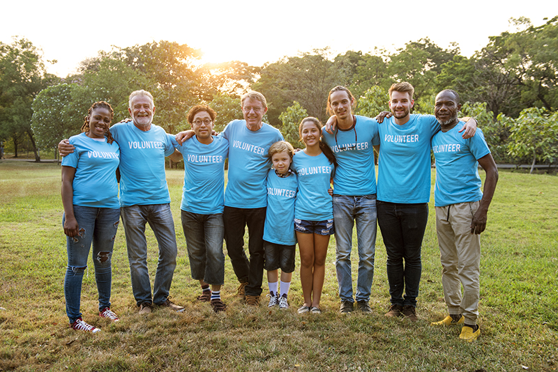 Group of adults and children wearing blue t-shirts with the word "Volunteer" across the chest, standing in an open field with the sun rising through the trees in the background.