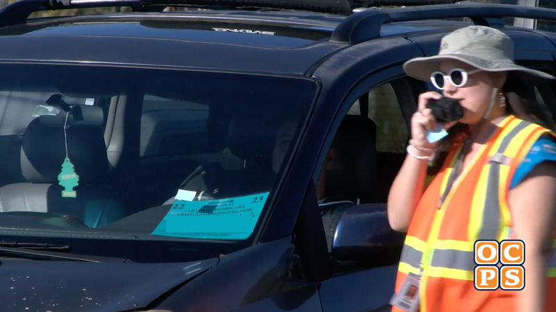 A female in a safety vest with a walkie directs a parent in a car 