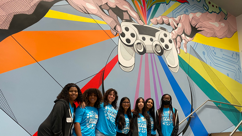 Seven girls stand in front of mural of video game controller