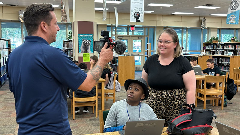 a man with a camera films a male student and female teacher