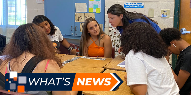 A teacher talks to a small group of learners with What's News logo in left corner