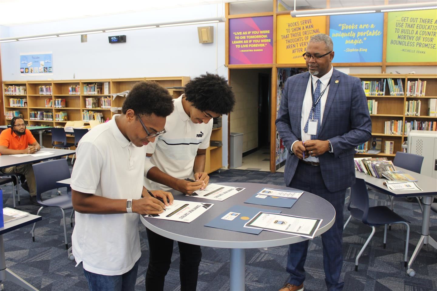 Two students sign forms as the superintendent looks on
