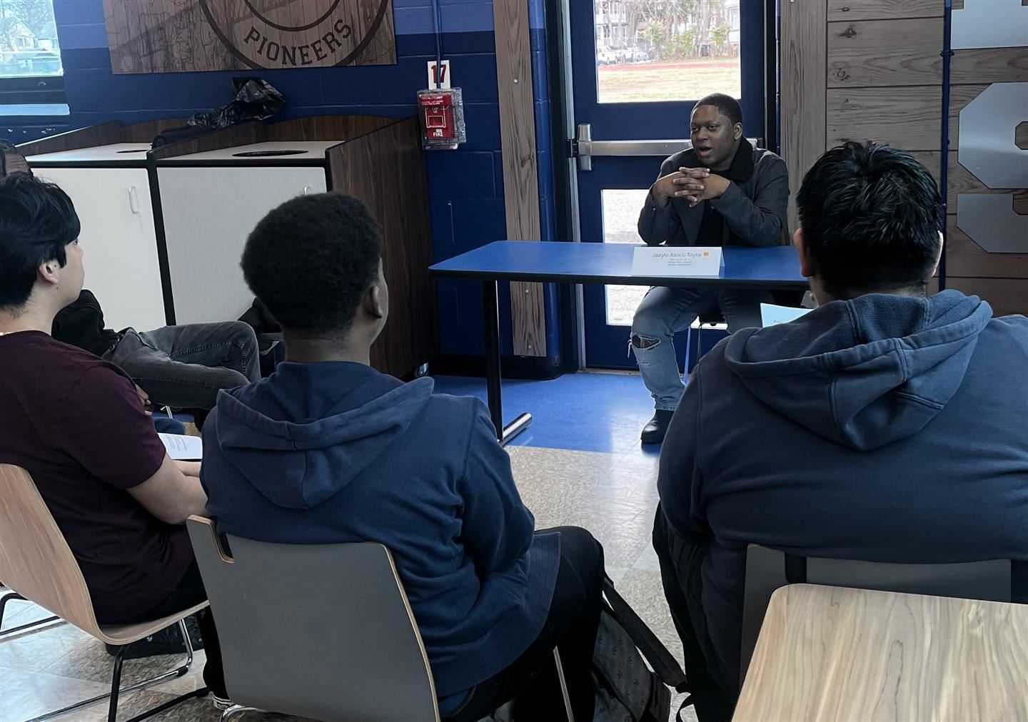 Three students sit in front of a table listening to a graduate speak