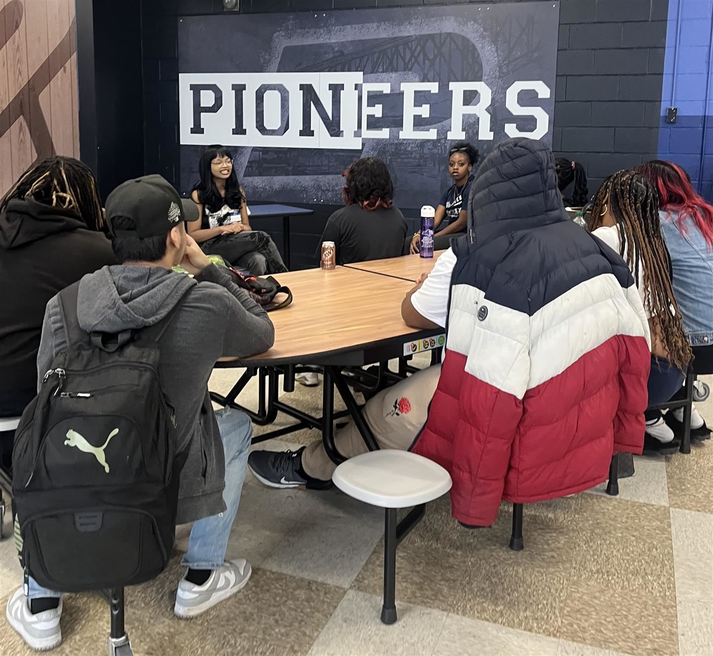  Students sit around a table listening to a graduate speak.