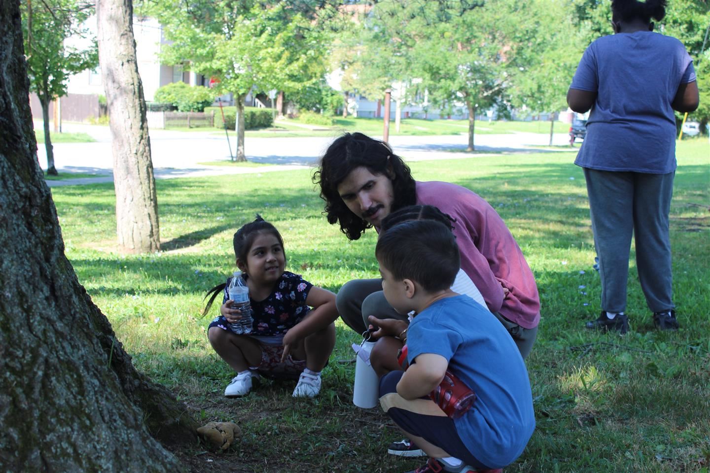 A teacher talks to two students while they squat near a tree