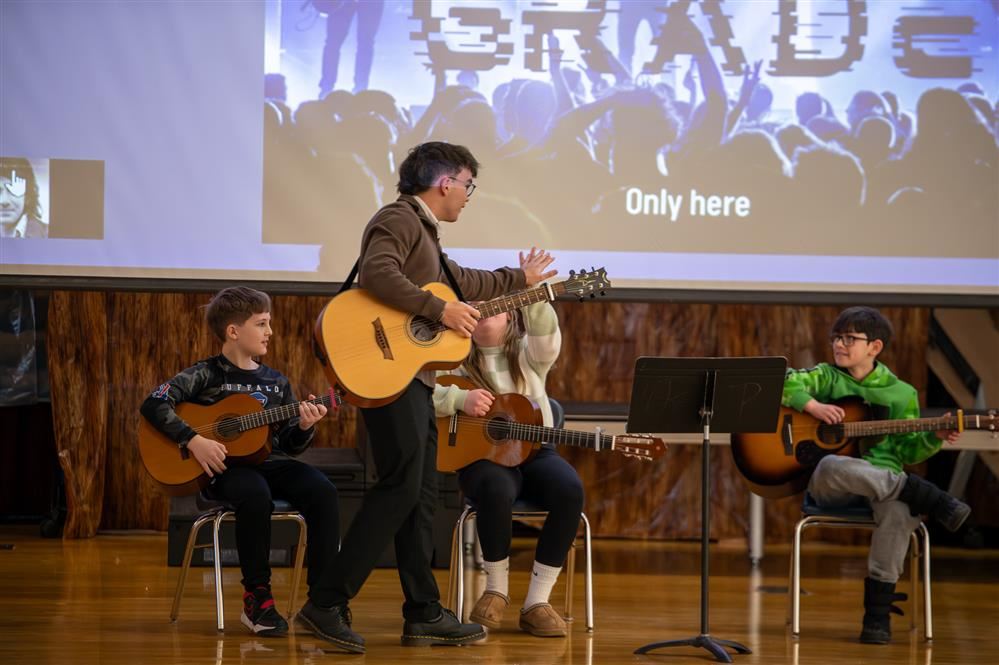 A guitar instructor stands in front of 4 seated students, high-fiving one after their performance.