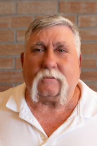 Headshot of Board Member Tony Bruno wearing white polo with brick background
