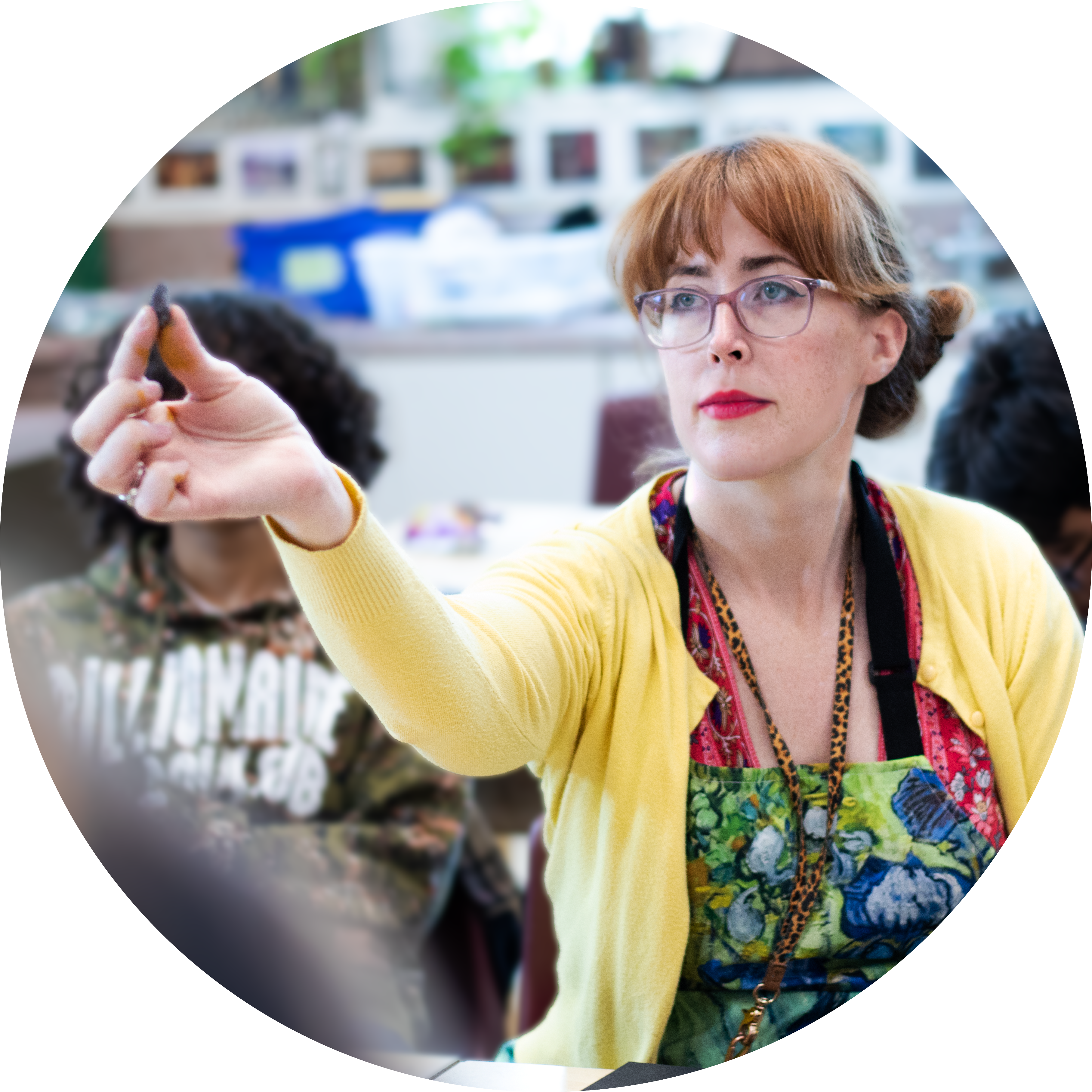 Female art teacher wearing yellow sweater holding pastel chalk in air to show students.