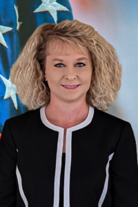 Profile picture of Governing Board Member Amy Petersmarck, caucasian female with blonde shoulder length wavy hair and blue eyes wearing a black suit coat with white trim. Background is American flag