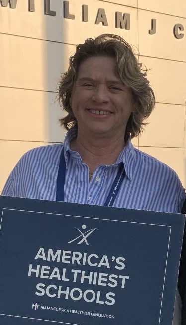 A woman with short, wavy blonde hair is smiling while holding a sign that reads "AMERICA'S HEALTHIEST SCHOOLS – A