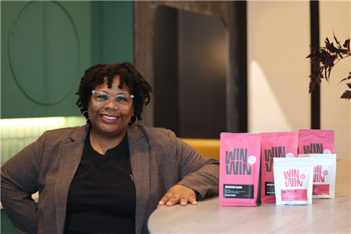A woman with short, curly hair and clear-framed glasses is smiling while leaning on a countertop. She is wearing a black shir