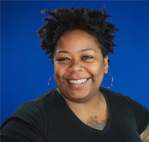 A woman with short, curly black hair is smiling against a solid blue background. She is wearing a black V-neck shirt, large g